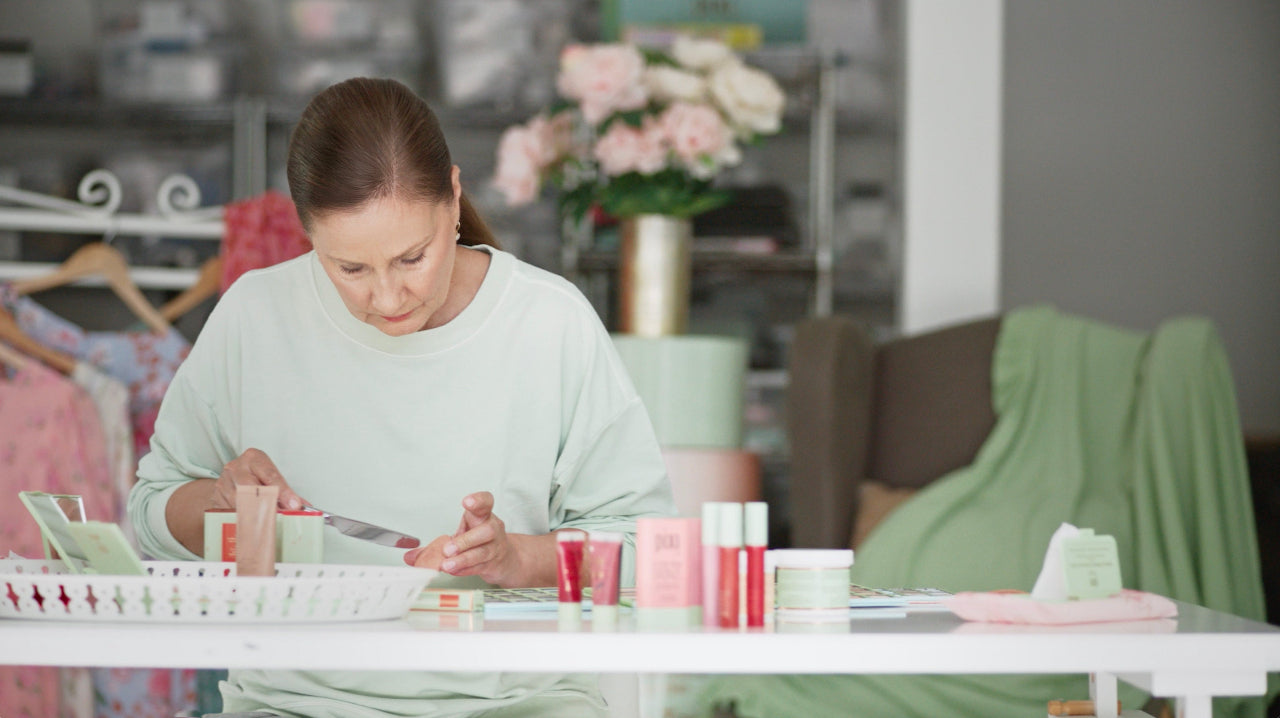 Petra in a light sage green sweatshirt sits at a white table, applying makeup from a small pink tube to her hand. Several cosmetic products in pink, red, and white packaging are arranged on the table in a softly lit room with a blurred background of clothing racks and flowers.
