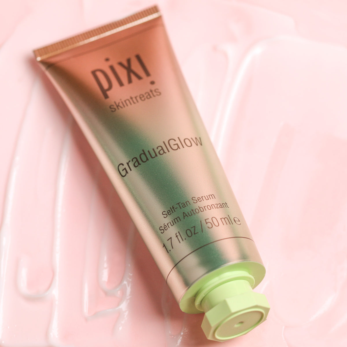 Pixi Skintreats GradualGlow Self-Tan Serum in a metallic rose gold tube with a light green cap, resting on a pink background with a smear of white serum visible.